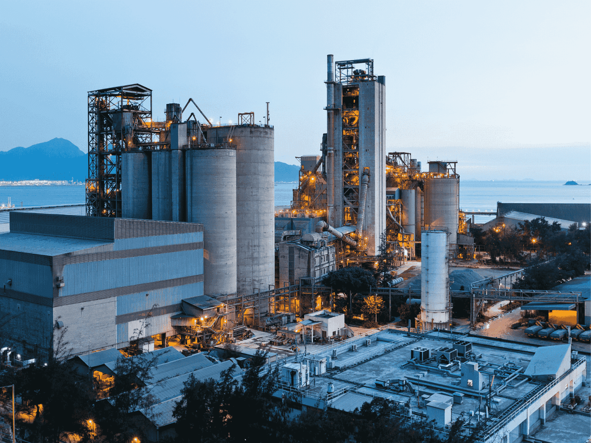 Industrial factory with large silos and machinery near a coastline, illuminated in the evening.